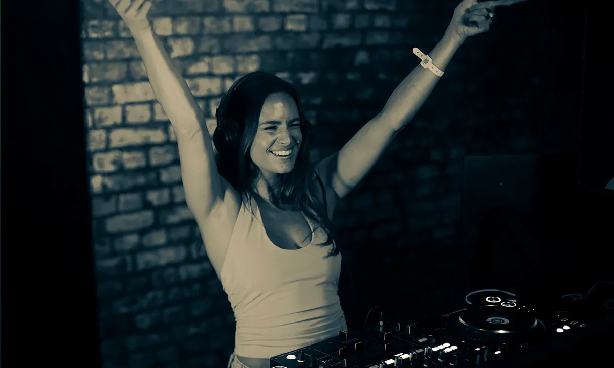 A woman DJ raises both hands high in the air and shows an elated expression