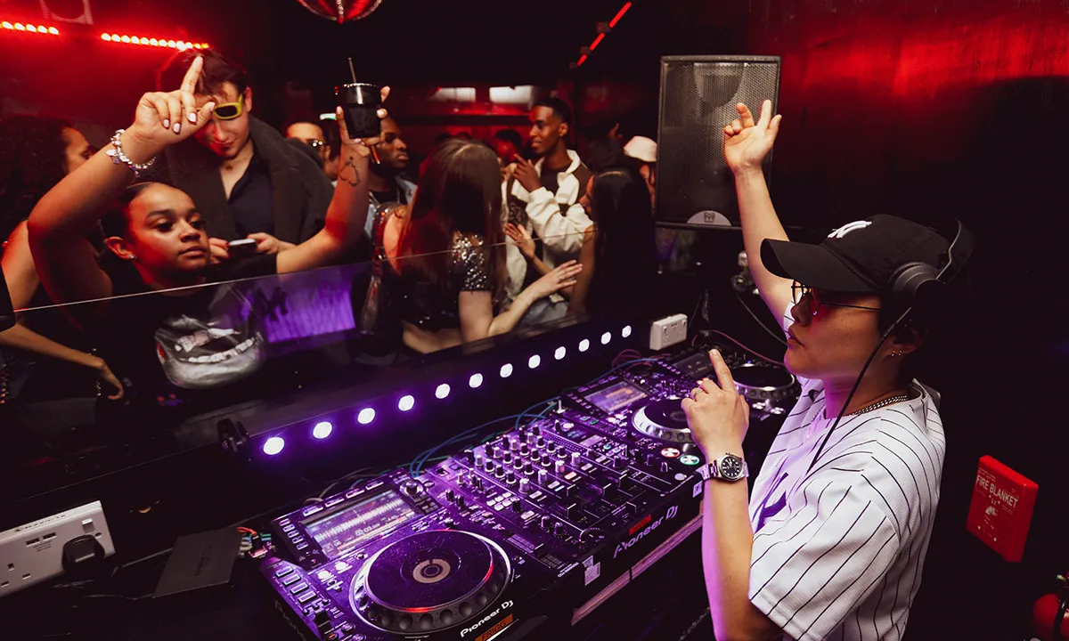 A female hip-hop DJ is performing to a busy room while her audience waves their hands