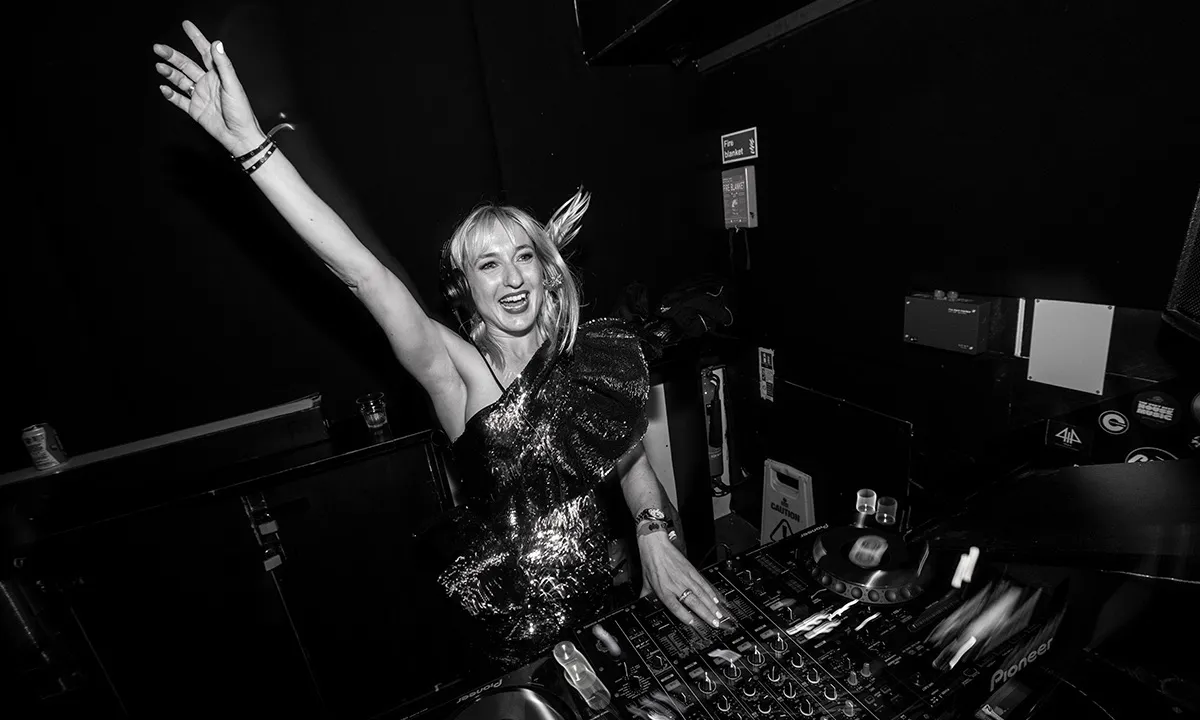 A female DJ raises her hand in the air and smiles while DJing