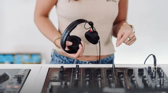 A woman holds DJ headphones in front of a DJ mixer