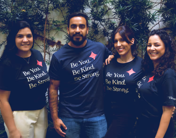 Quatro pessoas felizes usando camisetas pretas com as frases "Be You. Be Kind. Be Strong.", posando juntas ao ar livre diante de plantas verdes.