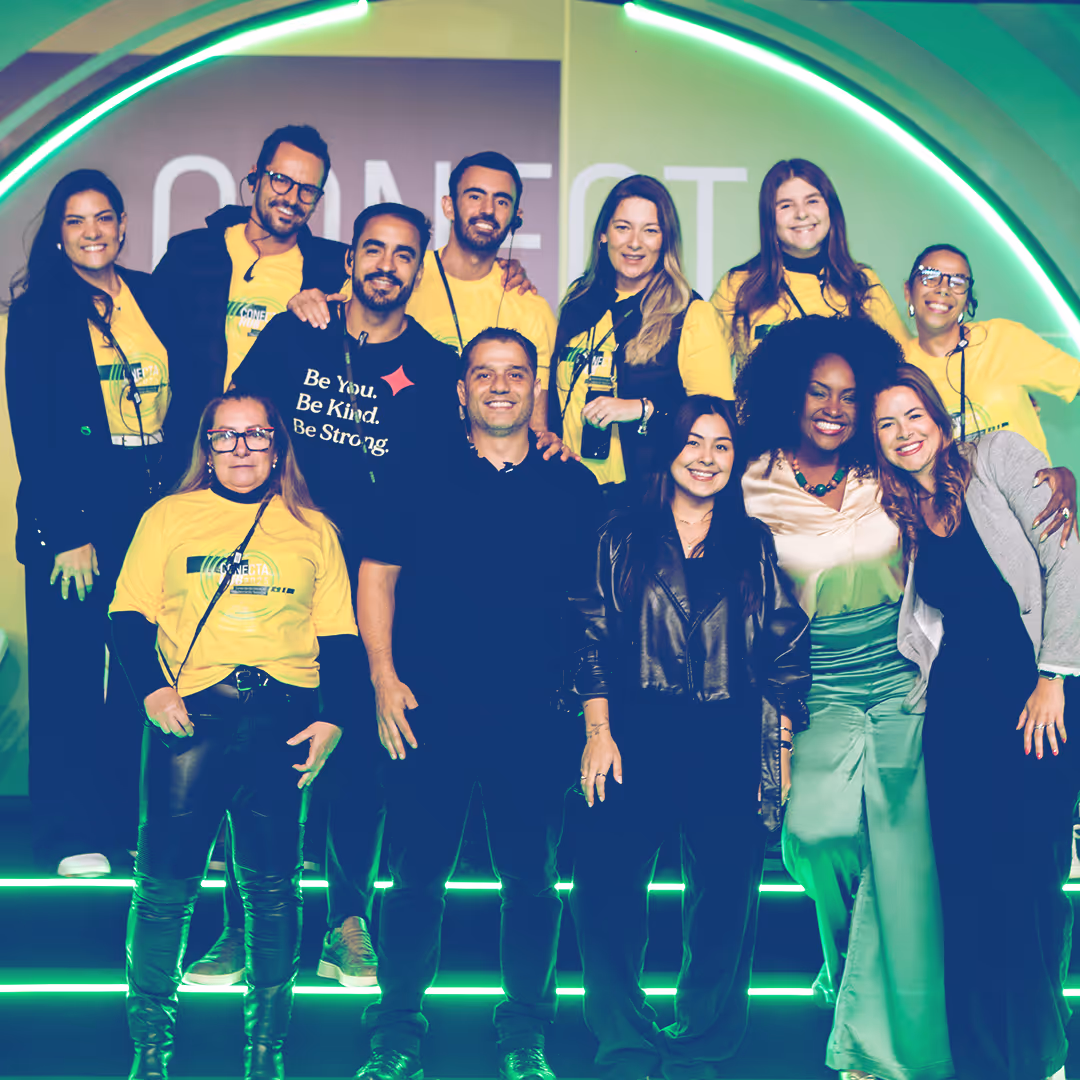 Grupo diversificado de 12 pessoas sorrindo em palco com iluminação verde e camiseta amarela, posando para foto em evento.