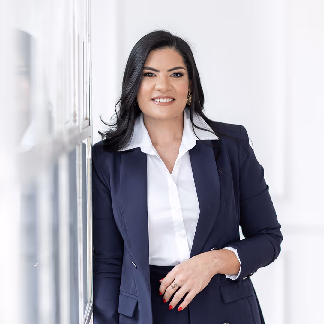 Mulher sorridente com cabelo preto, vestindo blazer azul escuro e camisa branca, encostada em uma parede branca.