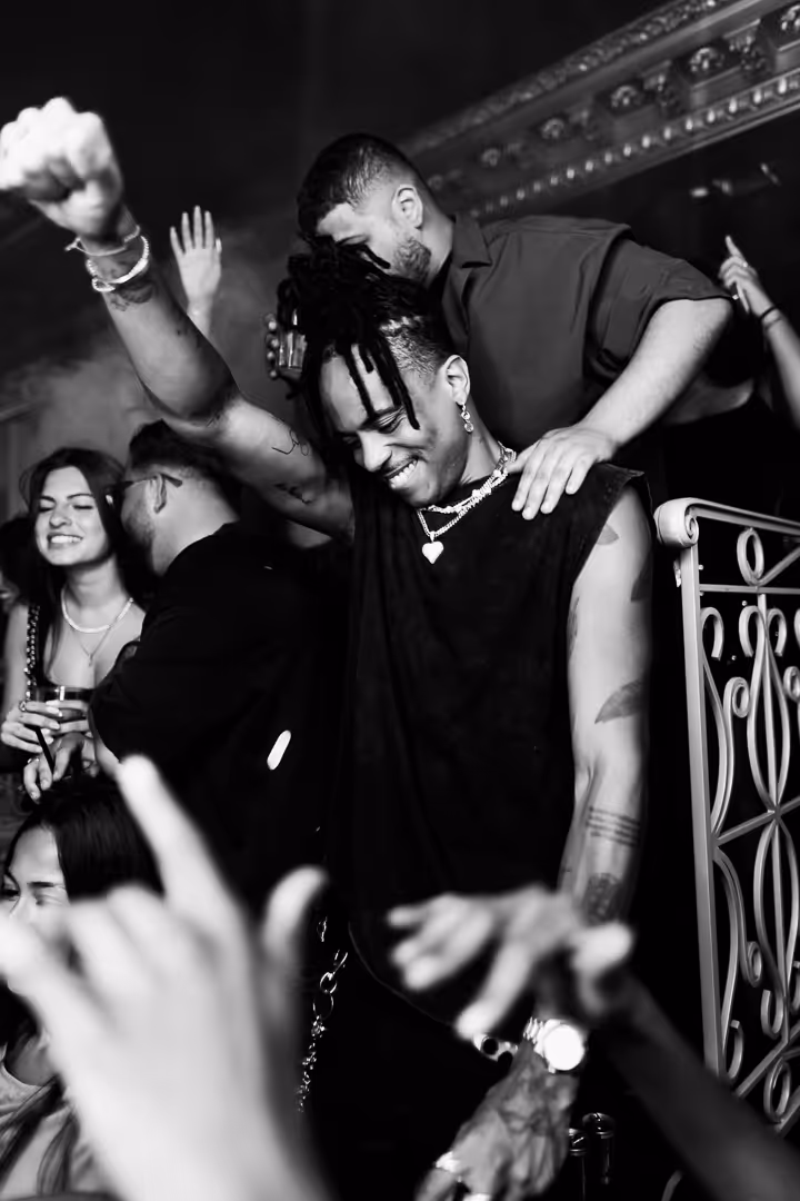 Black and white photo of a smiling man with dreadlocks raising his fist in celebration at a crowded party.