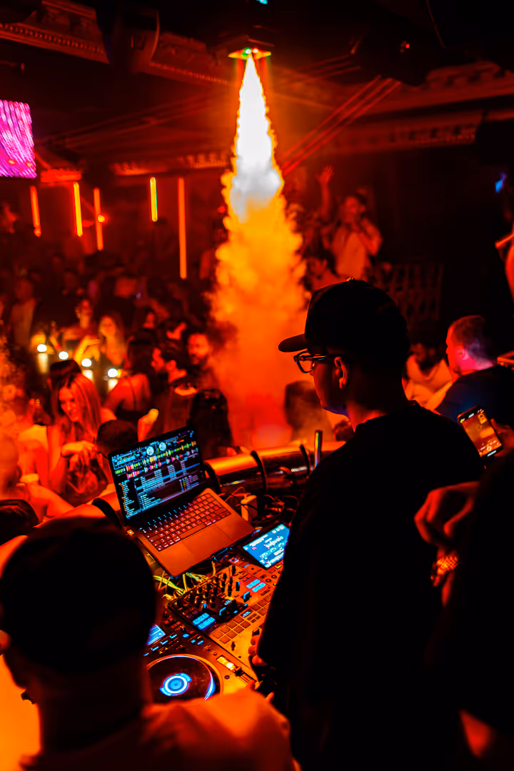 DJ performing at a crowded nightclub with laptop and mixing equipment, with bright orange smoke effects shooting upwards.