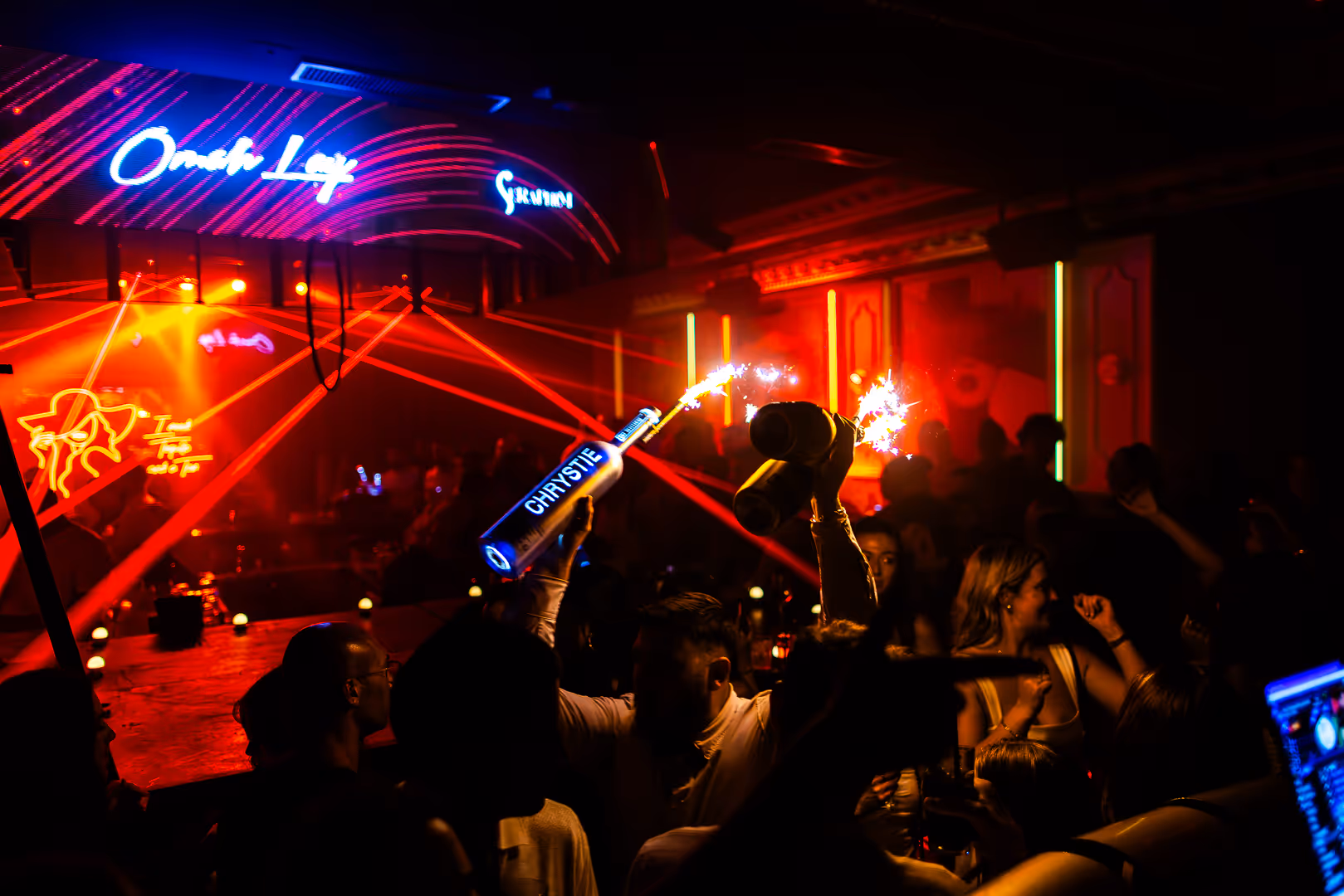 Crowded nightclub with people celebrating, illuminated by red laser lights and holding bottles with sparklers.