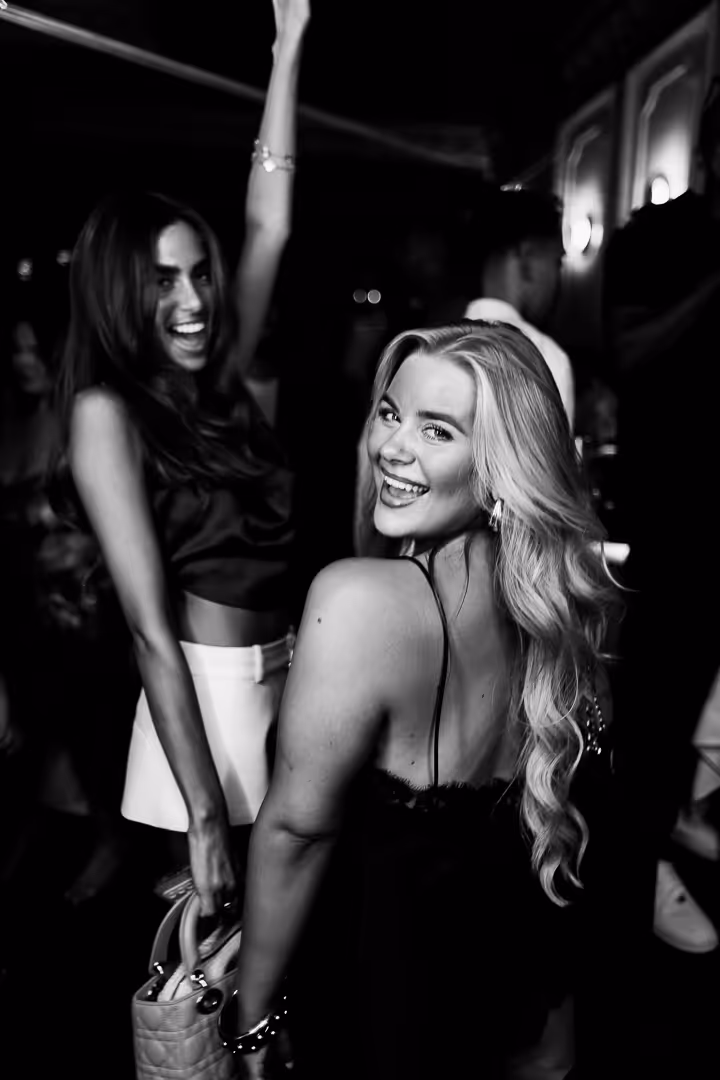 Two smiling women enjoying a night out, one raising her arm and the other looking back with long wavy hair.