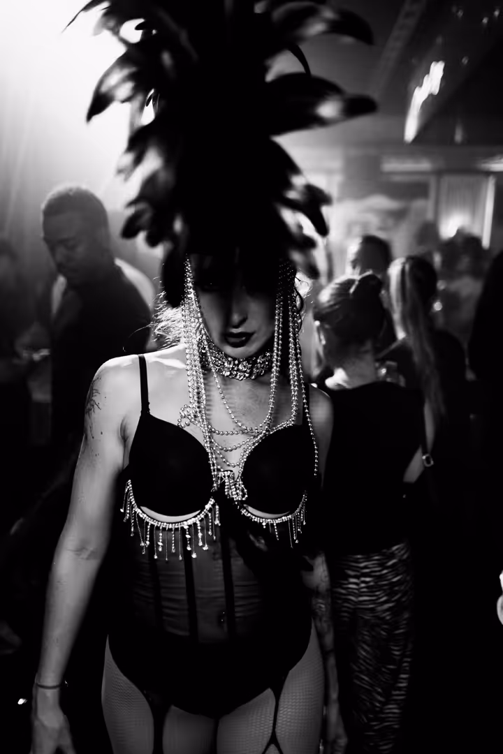 Person wearing elaborate headdress with feathers and pearl necklaces, dressed in black lingerie with beaded fringe, in a dark club setting.