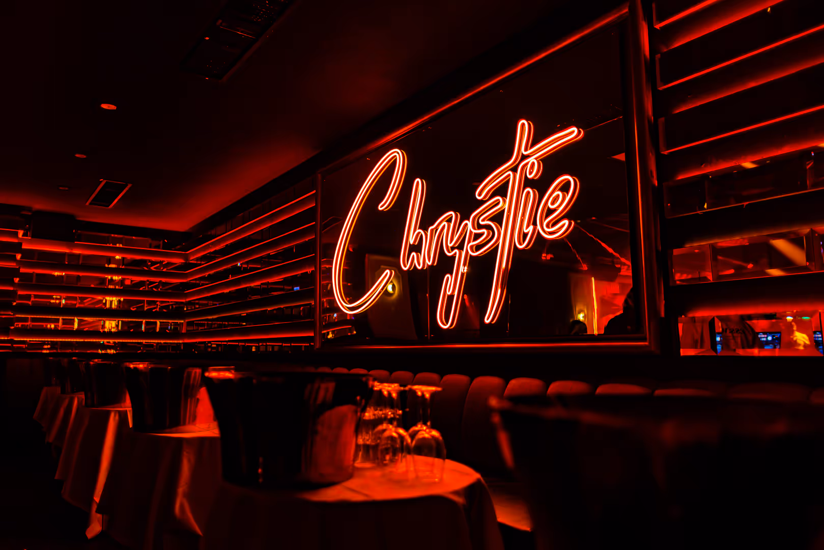 Interior of a dimly lit bar or lounge with red neon lights spelling 'Chrystie' on the wall and tables set with upside-down wine glasses and ice buckets.