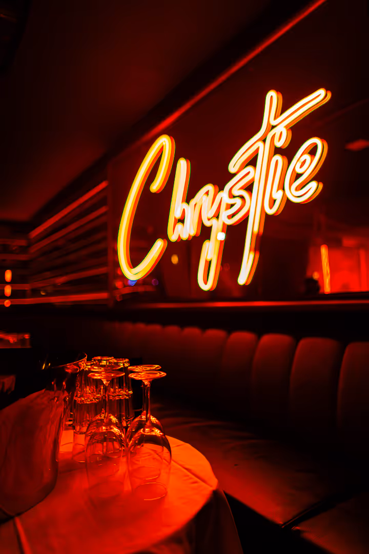 Dimly lit bar interior with a glowing neon sign reading 'Christie' and stacked upside-down wine glasses on a table.