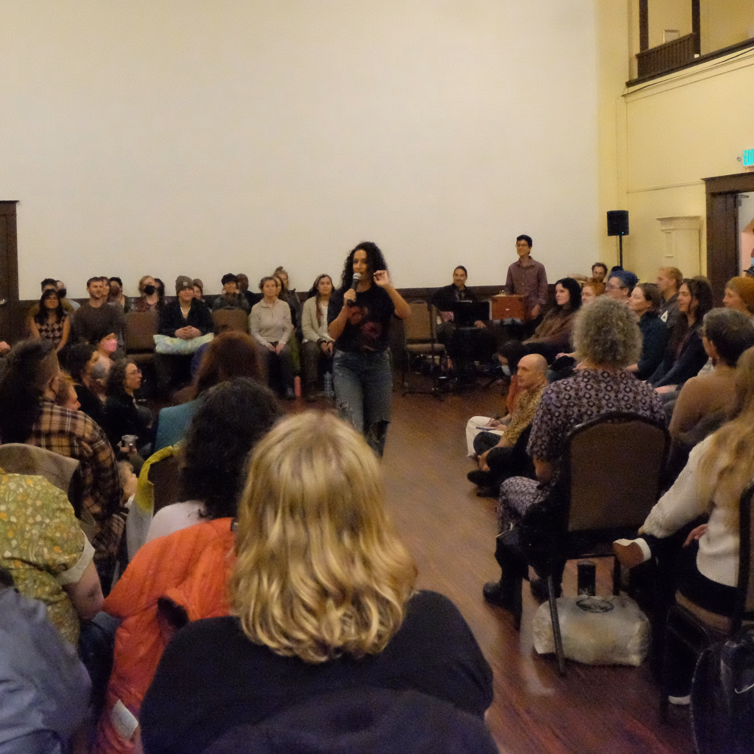 Lydia stands in the middle of a room speaking to an attentive audience seated around her in chairs and on the floor in a large room with wooden flooring.