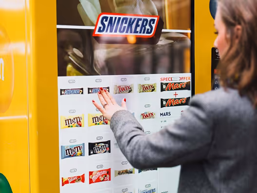 Woman selecting a chocolate bar from a branded Snickers vending machine with various candy options.