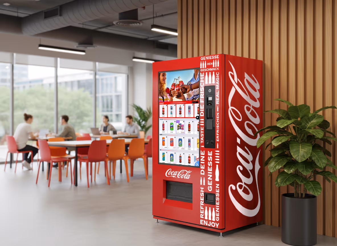 Red Coca-Cola vending machine with digital display in a modern office with people seated at a table in the background.