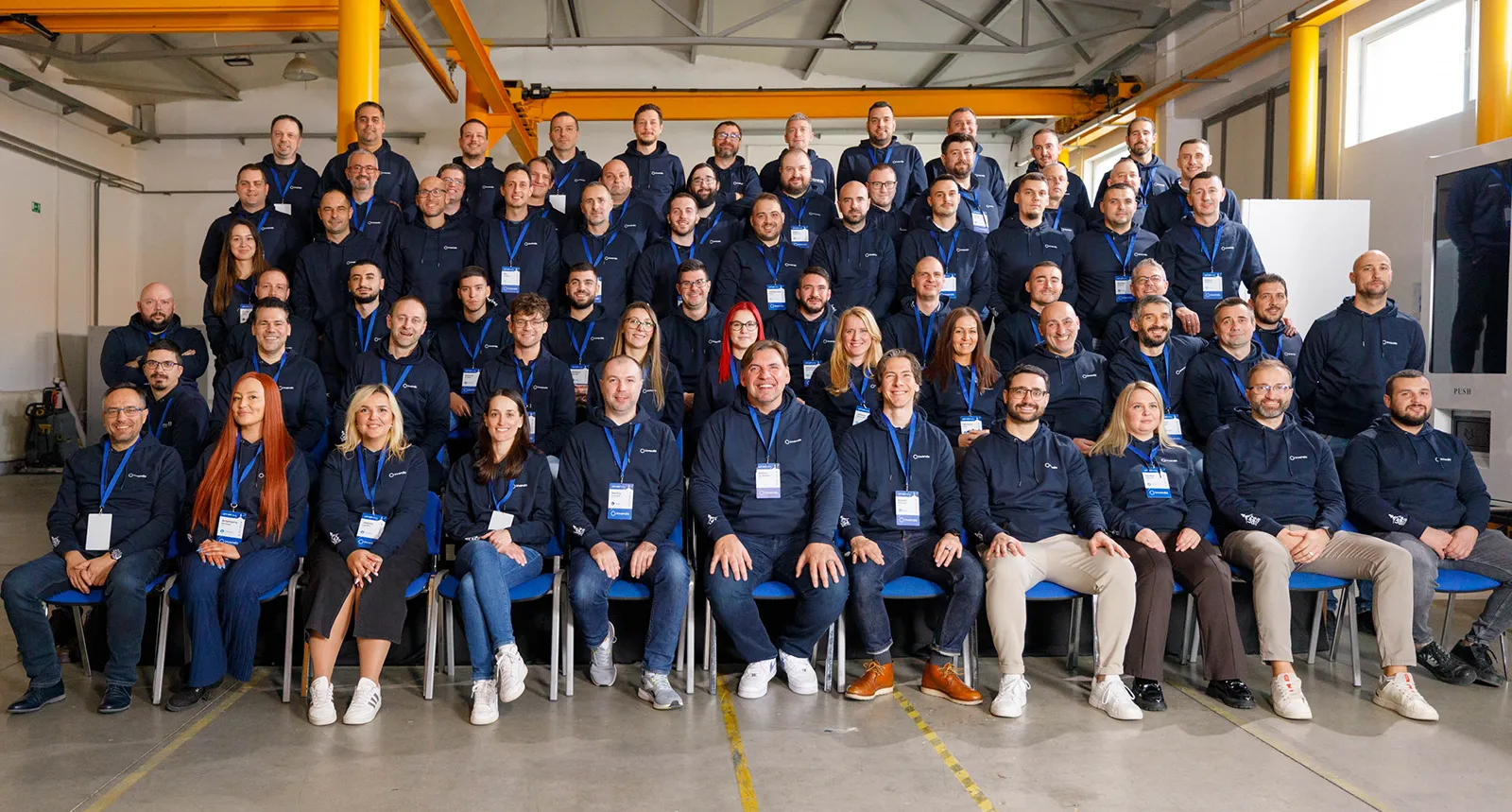 Large group of people wearing matching navy blue hoodies with name badges, seated and standing in rows inside a spacious industrial room.