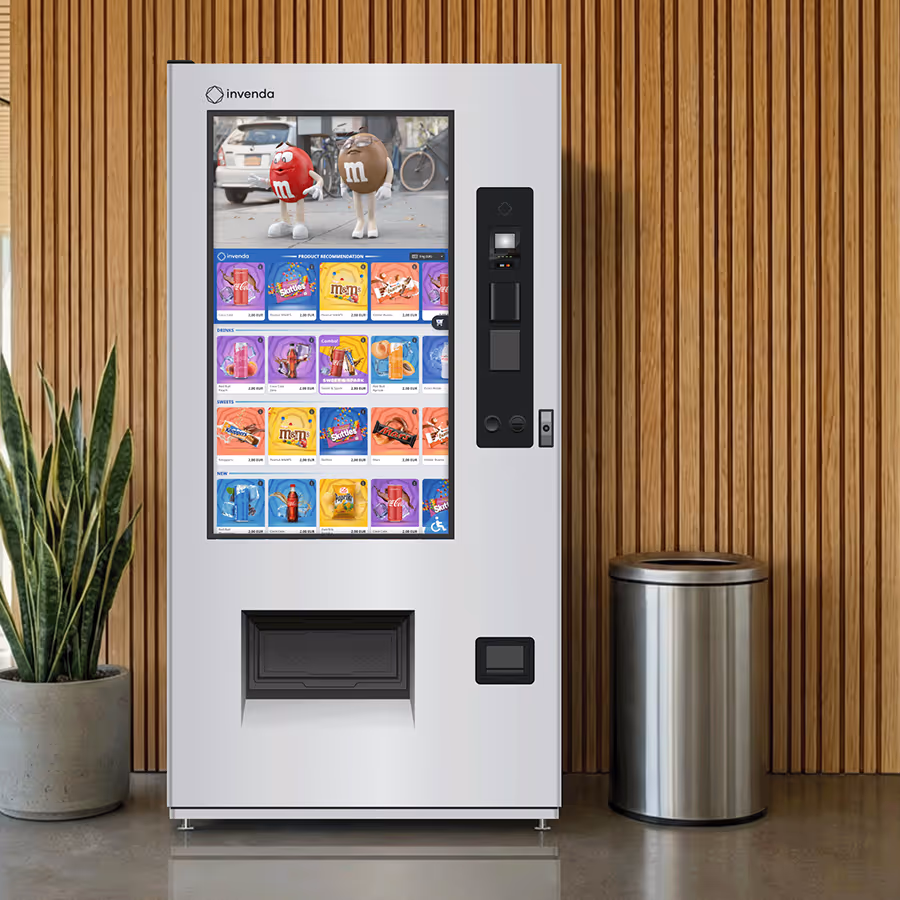 Modern white Invenda indoor vending machine with a digital touch screen displaying snacks, located next to a green plant and a stainless steel trash can against a wooden panel wall.