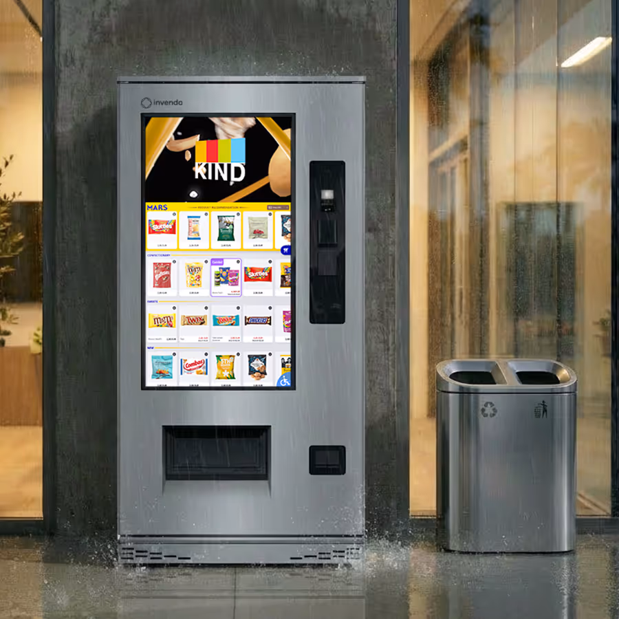Modern outdoor vending machine with digital touch screen displaying snack options, placed next to a dual compartment recycling and trash bin on a rainy day.