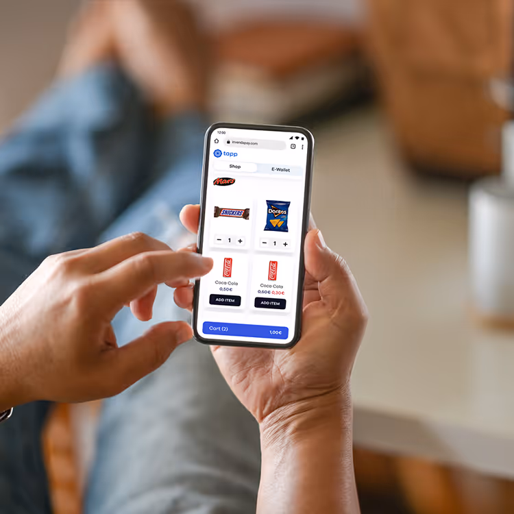 Person holding smartphone showing an app interface to shop snacks like Mars, Snickers, Doritos, and Coca Cola with prices and add item buttons.