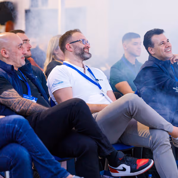 Group of men seated and laughing during an indoor event, some wearing lanyards and casual attire.