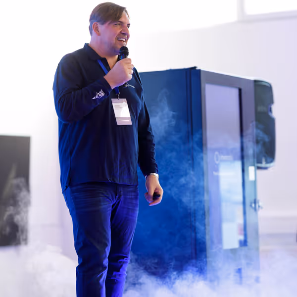 Man in a dark shirt speaking into a microphone on a stage with a Vending machine and smoke effects around him.
