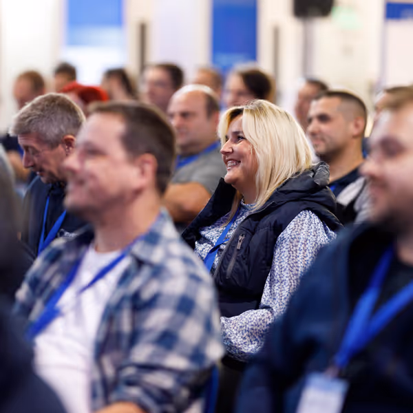 A group of adults attentively listening at a conference or seminar, with a smiling blonde woman in focus.
