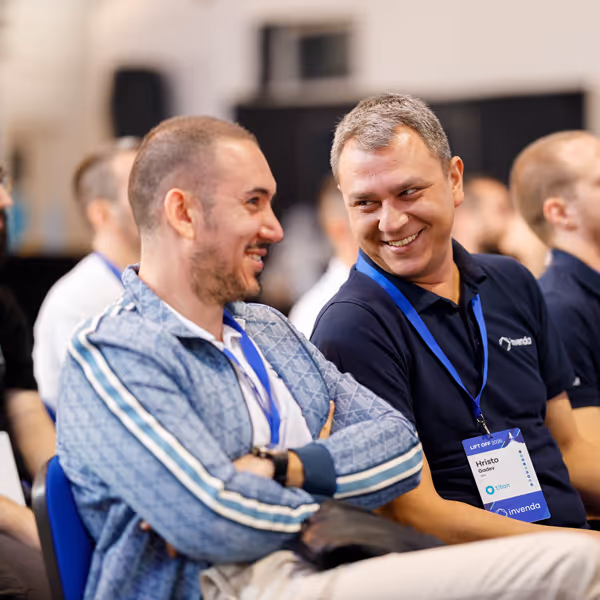 Two men sitting and smiling at each other during a conference or meeting, both wearing name badges and lanyards.