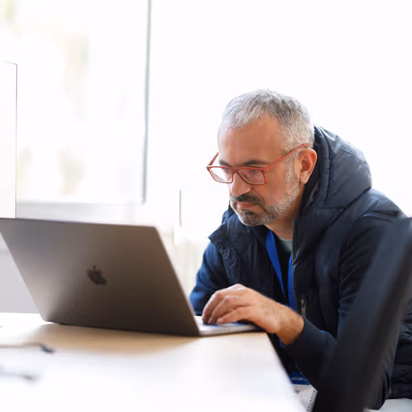 Man with gray hair and red glasses working intently on a laptop at a desk in a bright office.