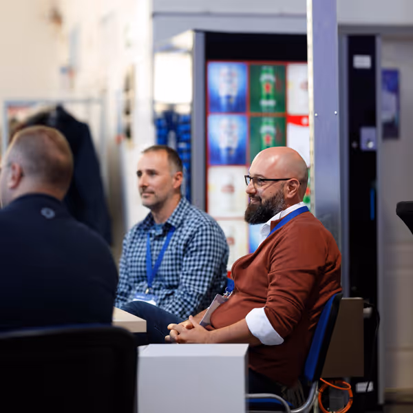 Two men sitting and attentively listening in a casual meeting, one wearing glasses and a rust-colored sweater. Vending machines blurred in the background.