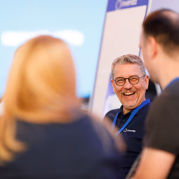 Smiling man wearing glasses and a navy blue shirt with an Invenda logo, engaging with two other people in a bright setting.