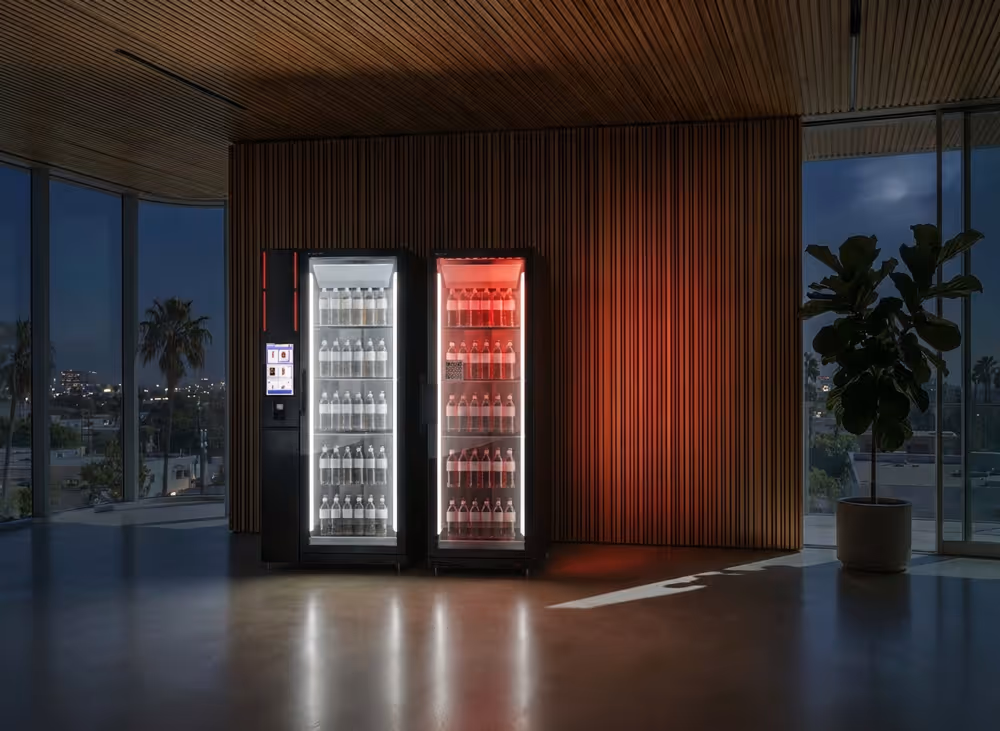 Two modern smart refrigerators stocked with bottled drinks against a wooden panel wall in a dimly lit room with large windows and a potted plant.