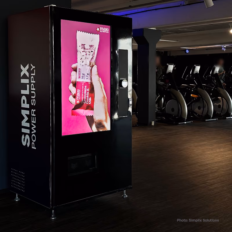 Man and woman interacting with a digital vending machine screen displaying snacks and drinks, with an 'Advertise here' space at the top.