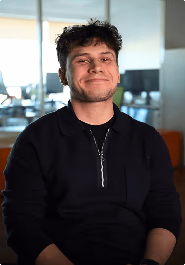 Jeune homme souriant portant un sweat noir avec une fermeture éclair, assis dans un bureau moderne.