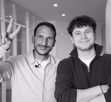 Deux hommes souriants debout dans un couloir, l'un fait un signe de paix avec ses doigts.