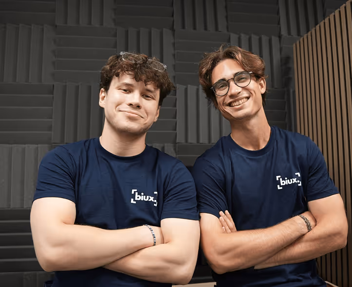 Deux jeunes hommes souriants portant des t-shirts bleus marine avec le logo 'biux' et les bras croisés devant un mur avec des panneaux acoustiques.