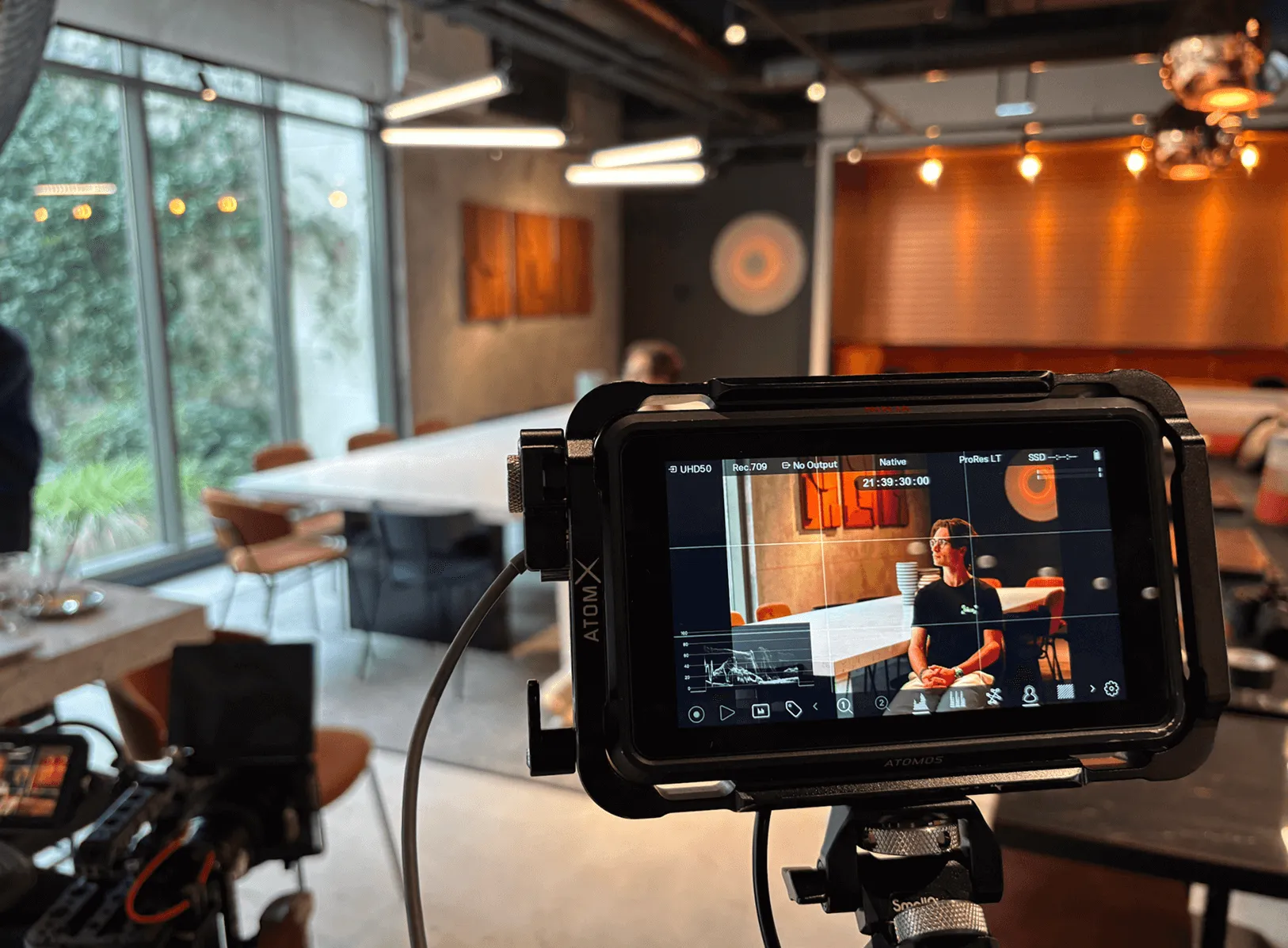 Écran de caméra montrant un homme assis dans une salle de réunion moderne avec une grande table blanche et des chaises orange.
