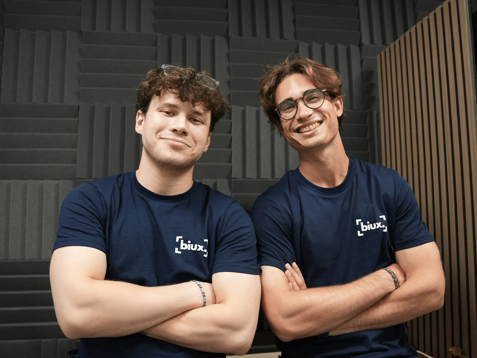 Deux jeunes hommes souriants avec des lunettes, bras croisés, portant des t-shirts bleu marine avec le logo Biux, devant un mur avec des panneaux acoustiques.