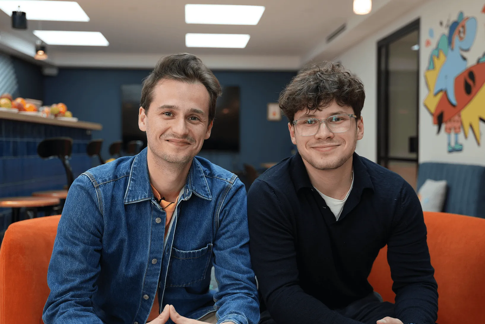 Deux jeunes hommes souriants assis sur un canapé orange dans un bureau moderne avec mur décoré et chaises en arrière-plan.