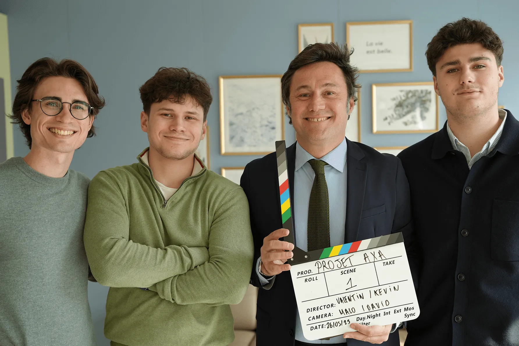 Quatre hommes souriants posant avec une claquette de cinéma portant l'inscription "PROJET AXA" et des détails sur la scène et les réalisateurs.