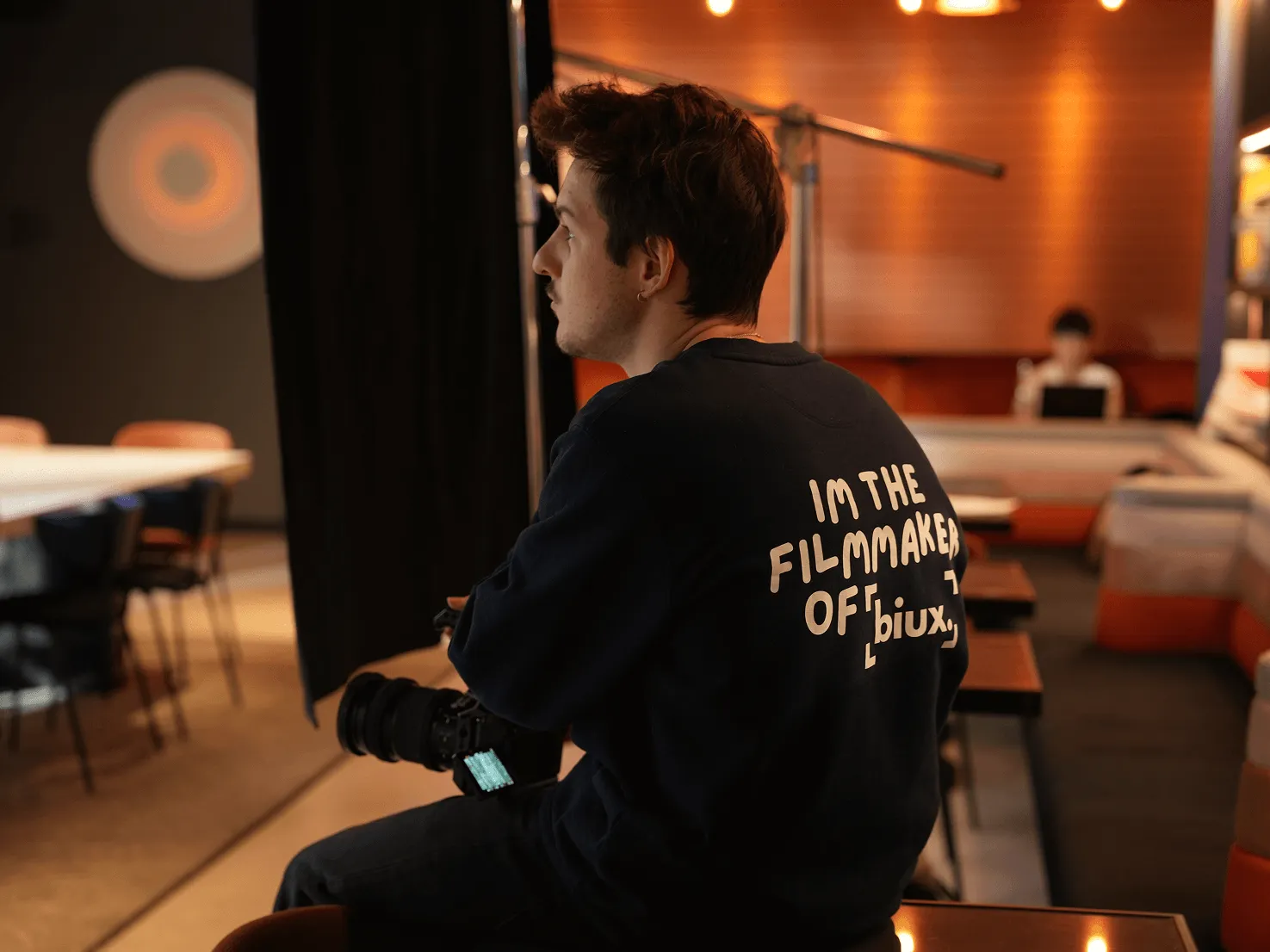 Homme assis tenant une caméra, portant un sweat-shirt avec l'inscription « I'm the filmmaker of biux » dans un intérieur chaleureux.