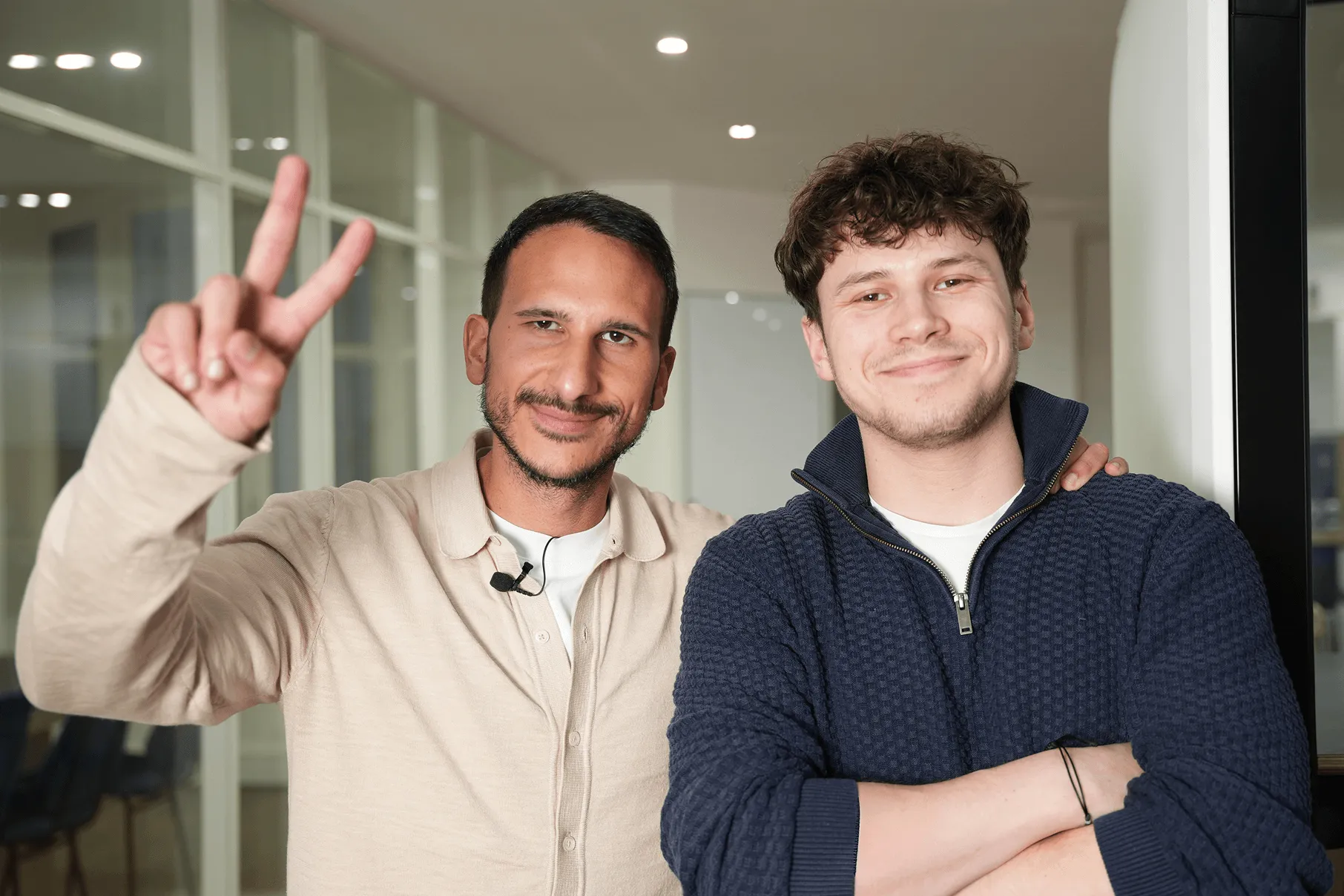 Deux hommes souriants posant ensemble dans un bureau, l'un fait le signe de la paix avec les doigts.