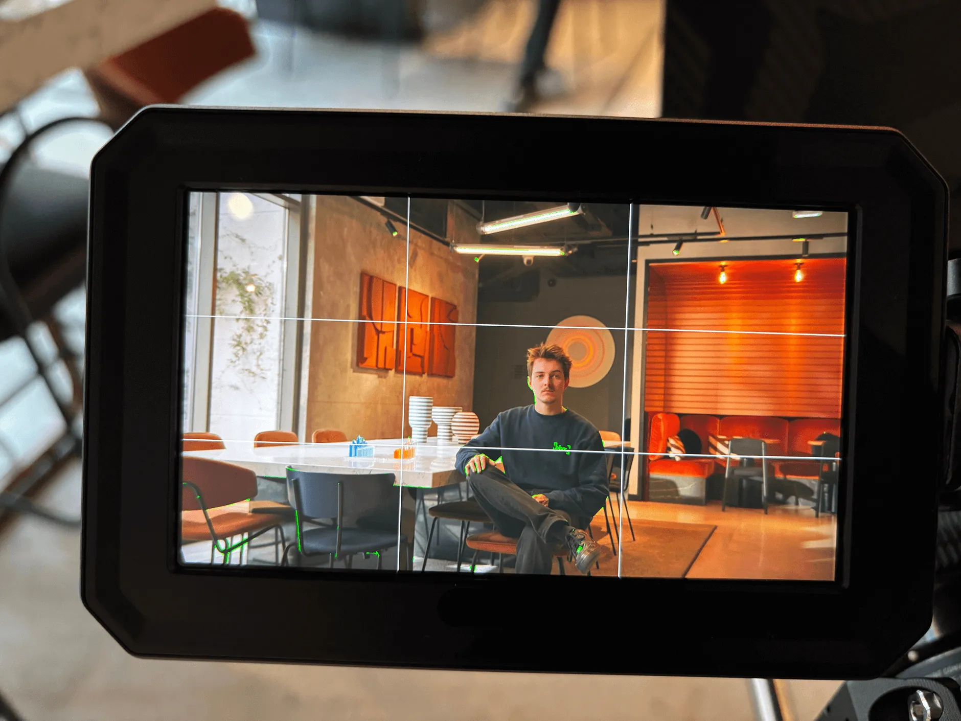 Un homme assis, jambes croisées, au centre d'une salle moderne avec table, chaises et décoration orange à l'arrière-plan.