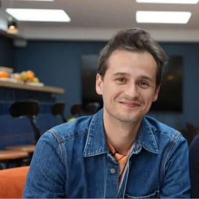 Jeune homme souriant portant une veste en jean dans un café avec des tabourets et un mur bleu en arrière-plan.