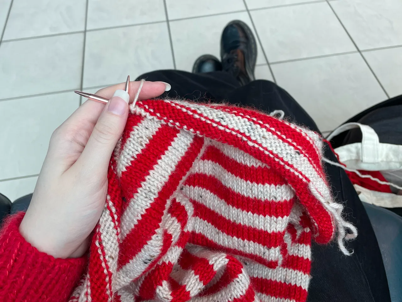 Person knitting a red and white striped fabric with metal knitting needles on tiled floor background.
