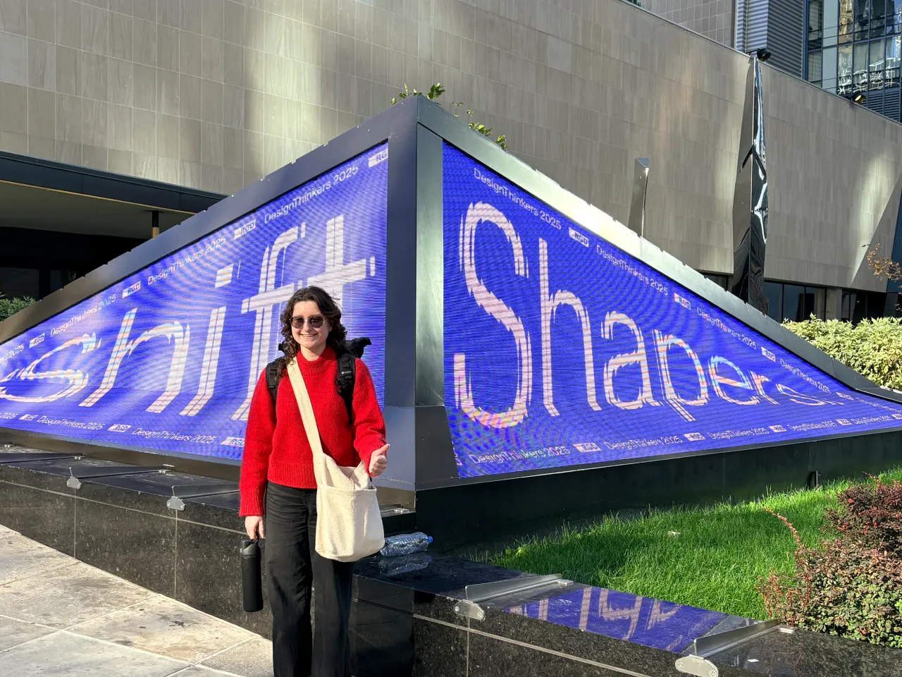 Person in a red sweater and sunglasses standing next to a large outdoor digital display showing the words 'Shift' and 'Shapers' in blue.