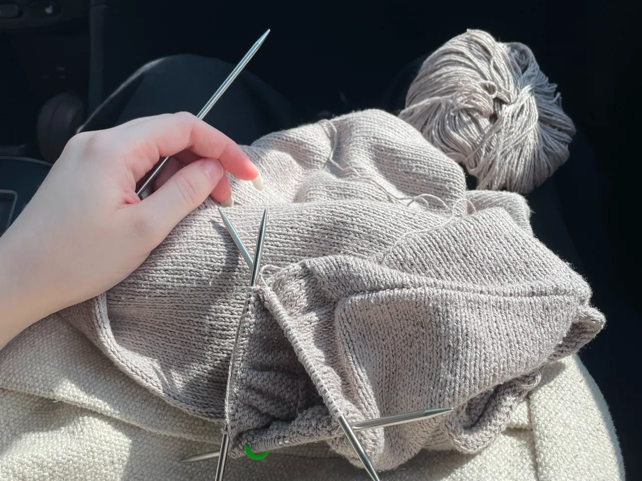 Hand holding knitting needles working on beige yarn with a ball of yarn in the background.