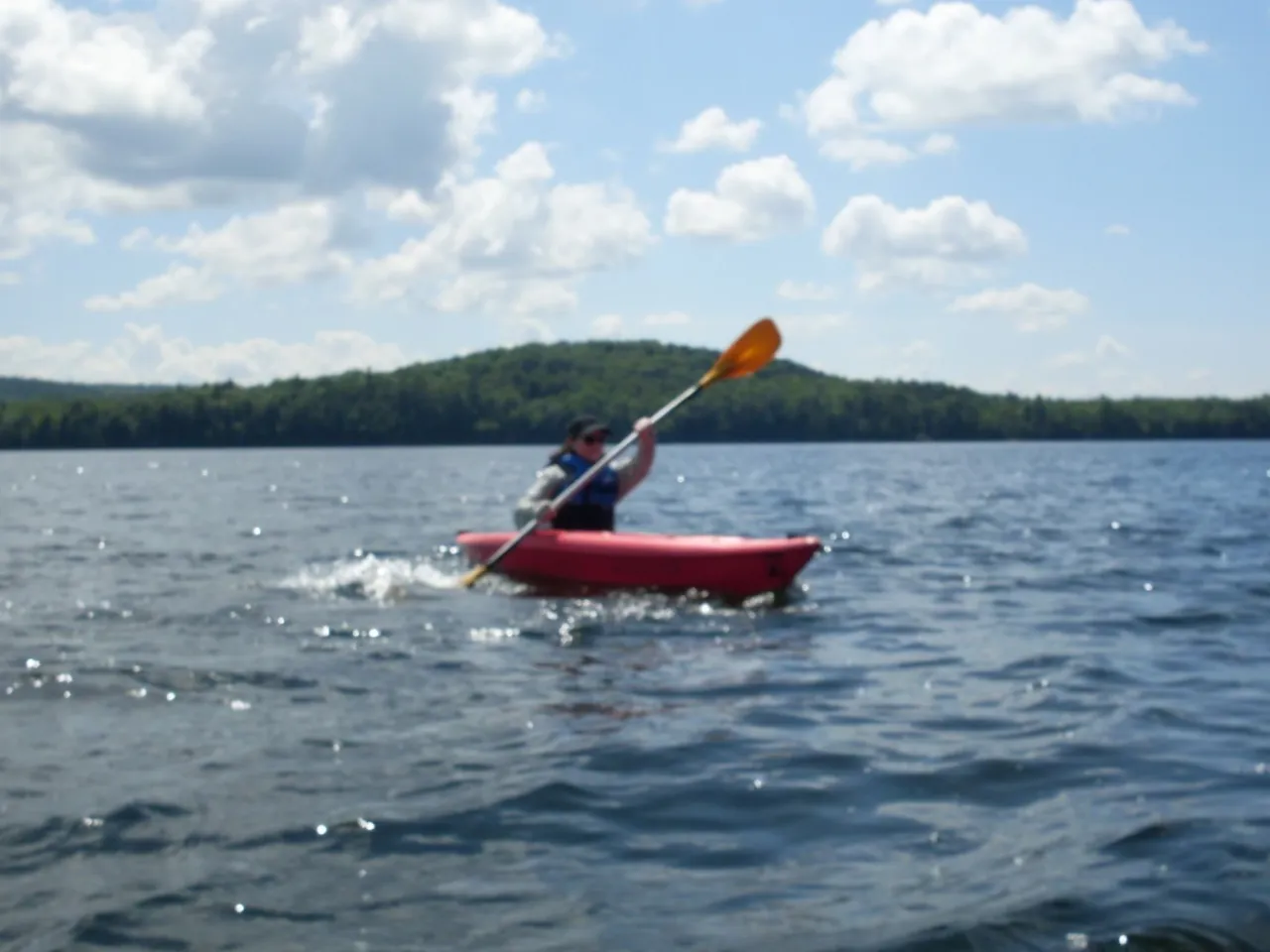 Person paddling a red kayak on a lake with a forested hill and partly cloudy sky in the background.