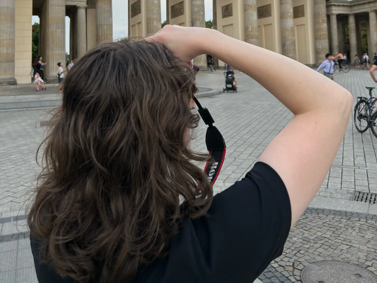 Person with long brown hair holding a camera strap preparing to take a photo in front of the Brandenburg Gate in Berlin.