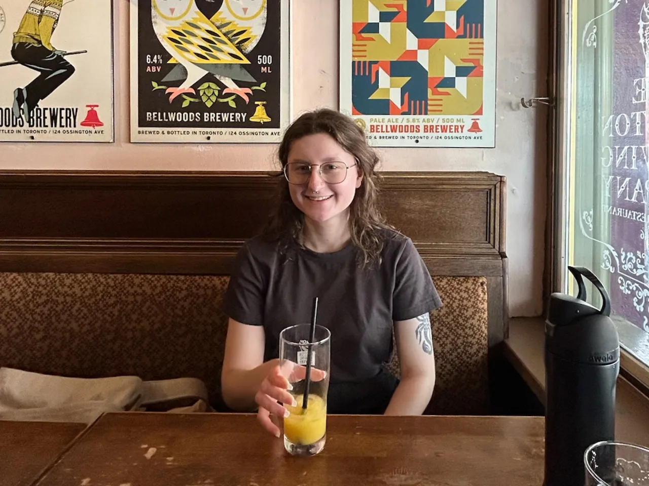 Smiling person with glasses and tattoo on arm sitting at a wooden table holding a glass with orange drink and black straw in a cozy restaurant booth.
