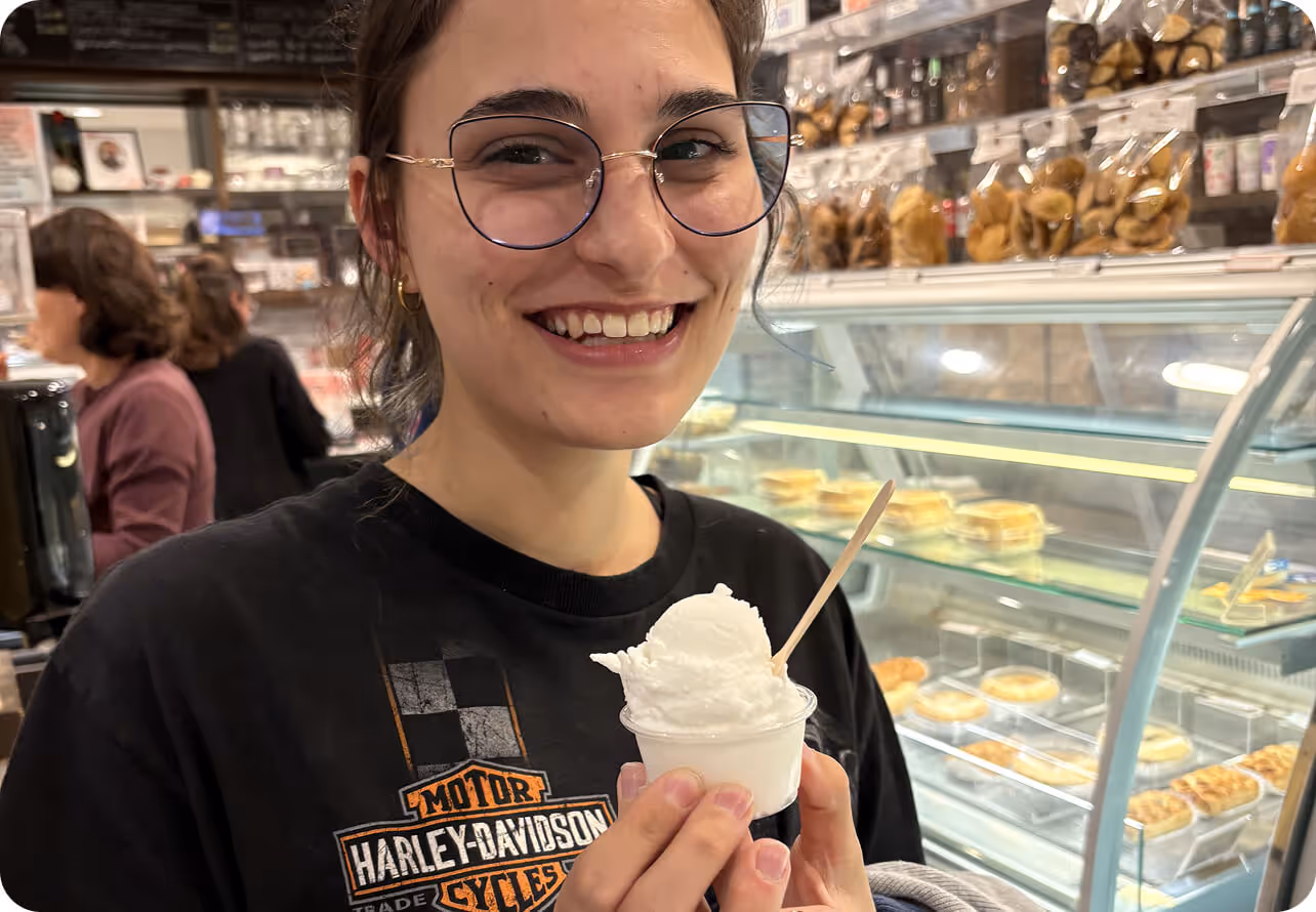 Smiling woman wearing glasses and a Harley-Davidson shirt holding a cup of white ice cream inside a café.