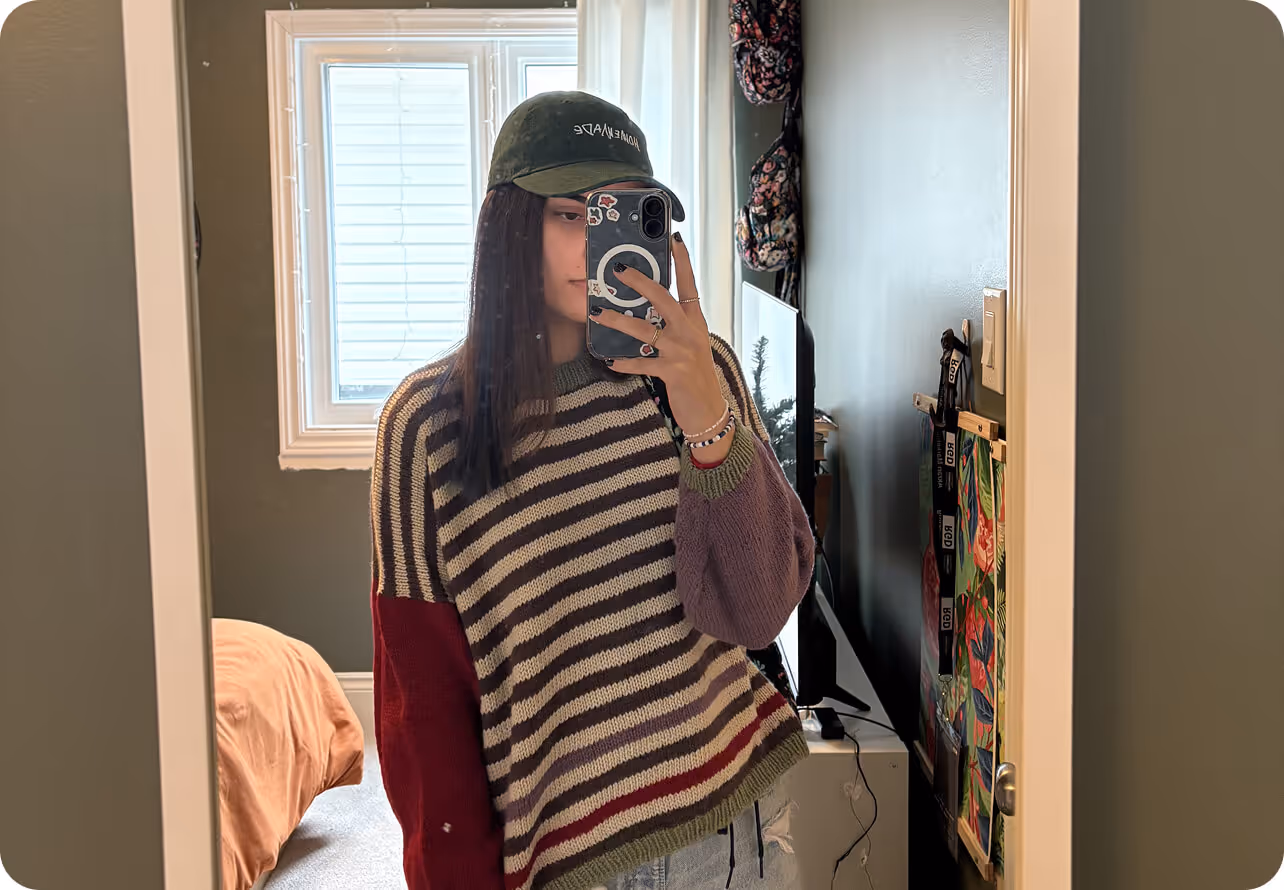 Person taking mirror selfie wearing a striped sweater with red and purple sleeves and a dark green cap.