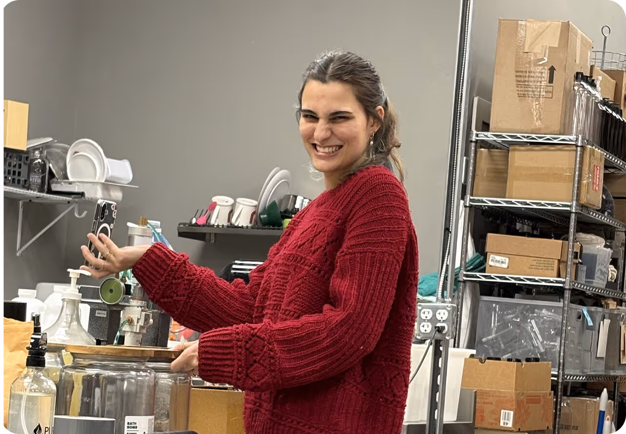 Smiling woman in a red sweater holding a phone, standing in a workspace with shelves of boxes and kitchenware.